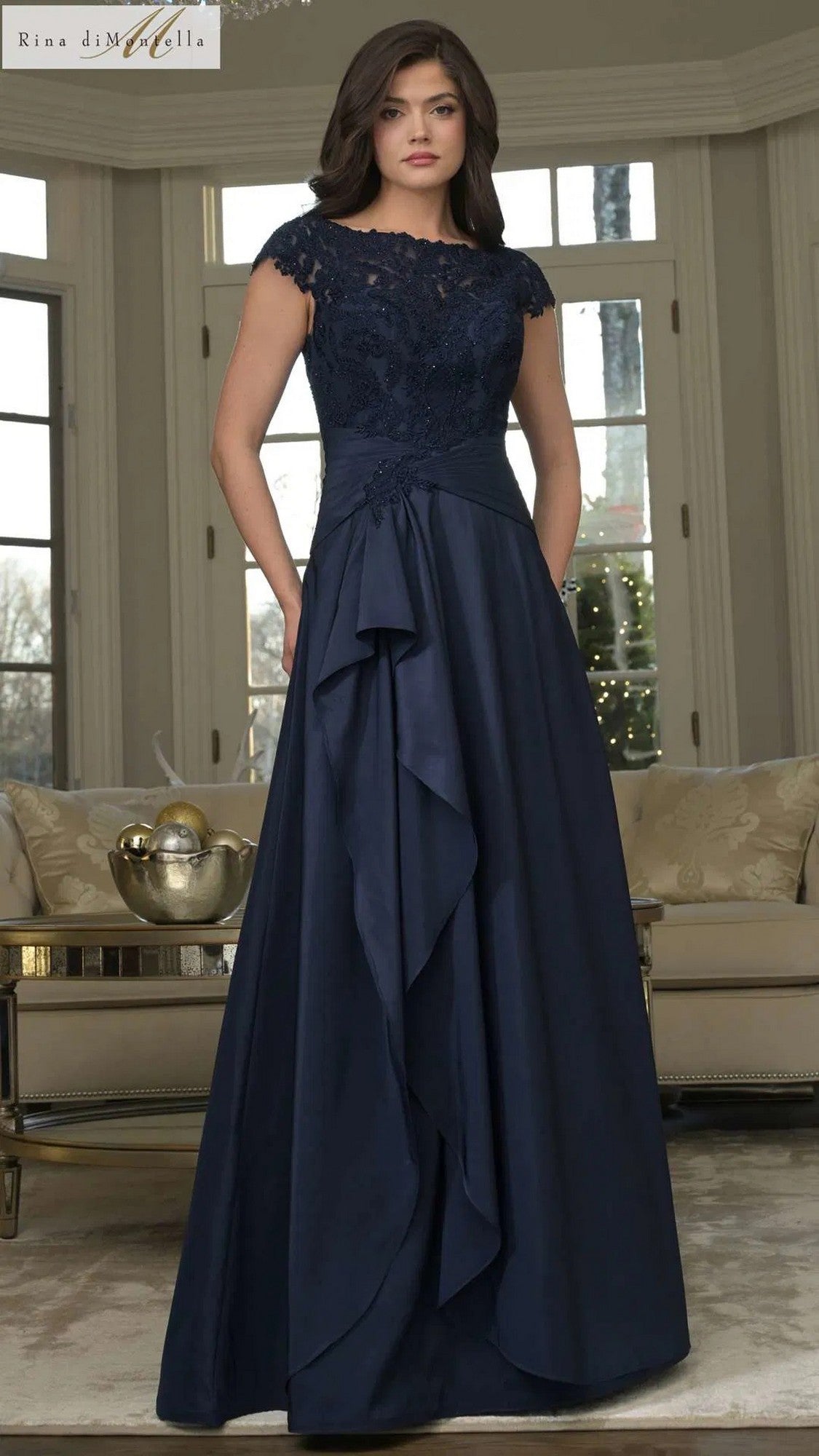 A woman stands indoors wearing the Rina Di Montella RD3327 long formal dress in navy, featuring a lace-embroidered bodice, cap sleeves, ruffled overlay, and a floor-length taffeta skirt.